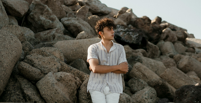 A man in a short-sleeved shirt sits on a rock by the sea and looks into the distance